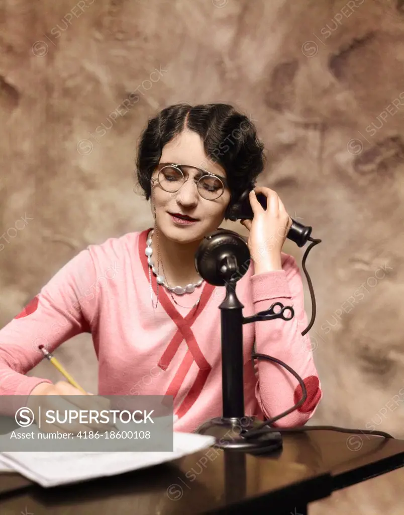 1920s WOMAN WEARING PINCE-NEZ GLASSES SITTING AT DESK TALKING ON CANDLESTICK PHONE AND WRITING