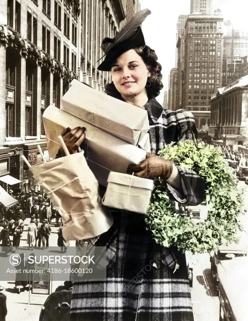 1930s 1940s WOMAN LOOKING AT CAMERA ARMS FULL WITH CHRISTMAS SHOPPING PACKAGES & WREATH COMPOSITE WITH STREET SCENE