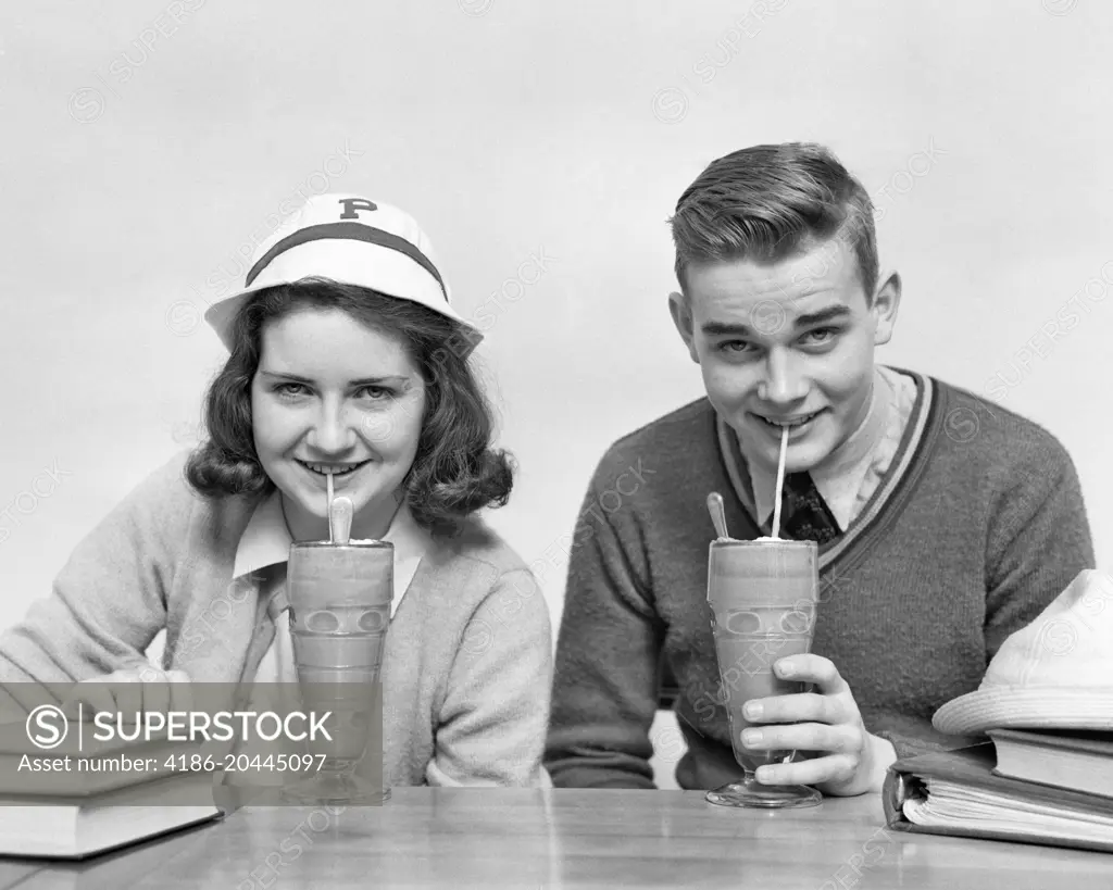 1940s TEENAGE BOY AND GIRL DRINKING MILKSHAKES TOGETHER LOOKING AT CAMERA