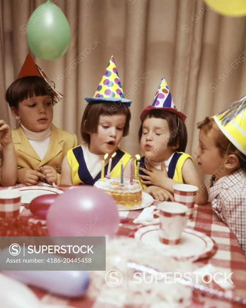 1970s TWIN GIRLS AND OTHER CHILDREN AT BIRTHDAY PARTY BLOWING OUT CANDLES ON CAKE