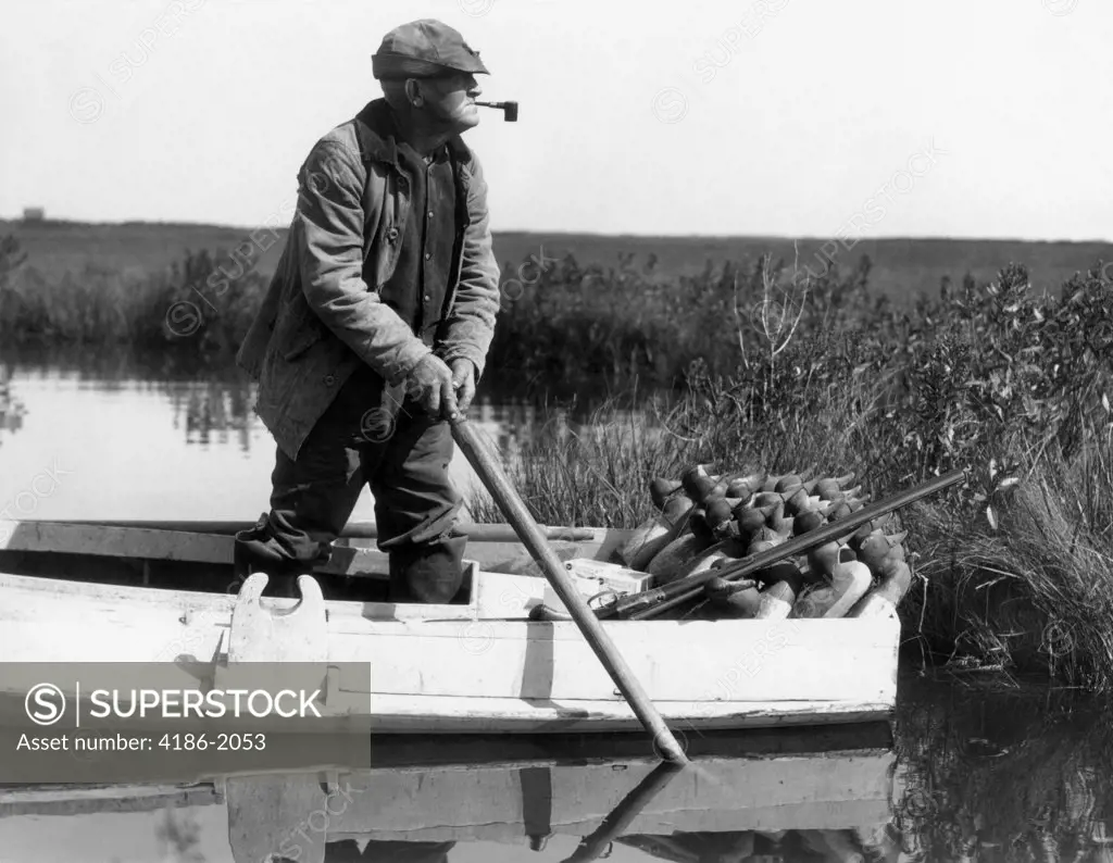1920S 1930S Senior Man Standing In Boat About To Deploy Duck Decoys In Wetlands Marsh