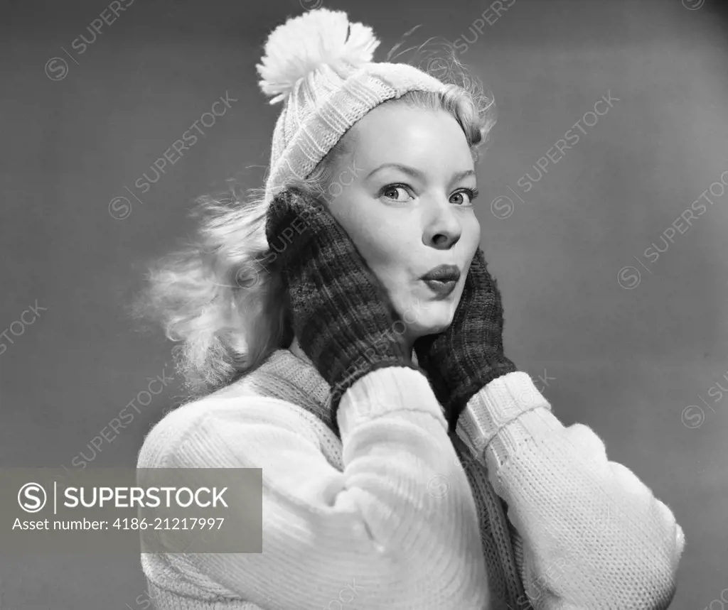 1950s YOUNG WOMAN PURSING LIPS HANDS COVERING EARS LOOKING AT CAMERA WEARING WINTER WOOL SWEATER MITTENS KNIT CAP