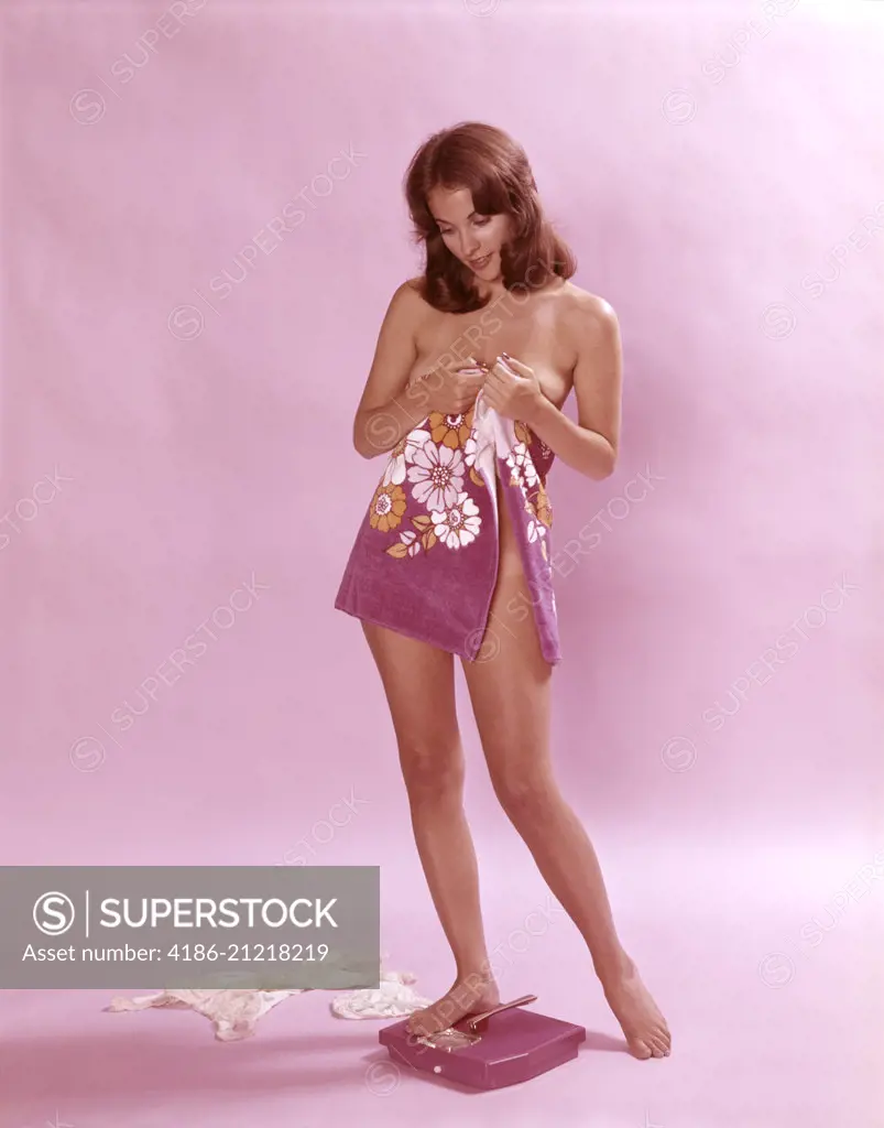 1970'S WOMAN WRAPPED IN TOWEL IN BATHROOM CHECKING WEIGHT ON SCALE INDOOR