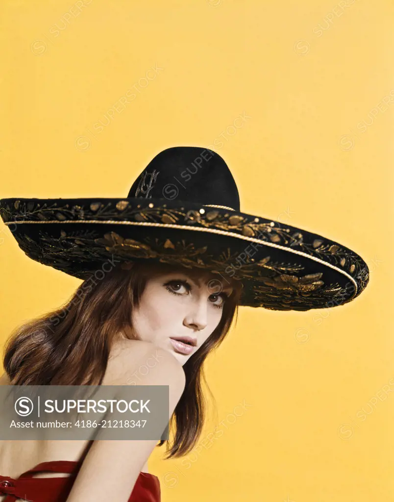 1970'S PORTRAIT ATTRACTIVE SULTRY BRUNETTE WOMAN WEARING GOLD EMBROIDERED BLACK MEXICAN SOMBRERO LOOKING AT CAMERA OVER SHOULDER