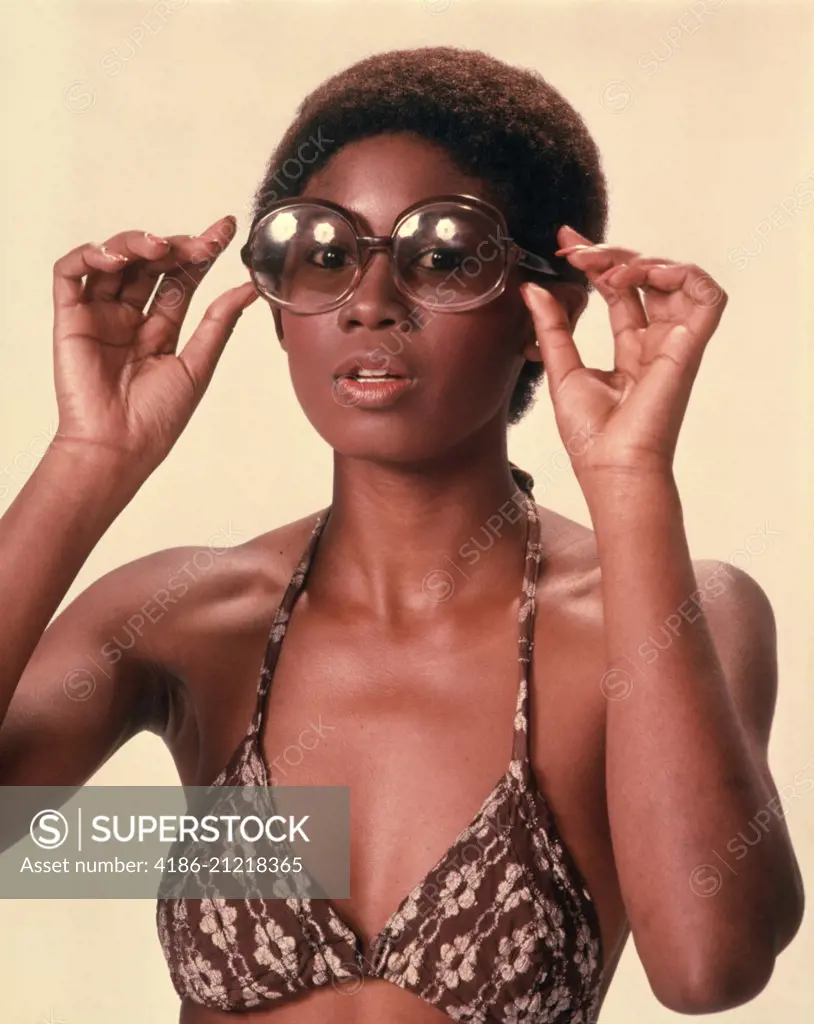 1970s AFRICAN AMERICAN WOMAN WEARING BIKINI BATHING SUIT TOP PUTTING ON LARGE LENS SUNGLASSES LOOKING AT CAMERA