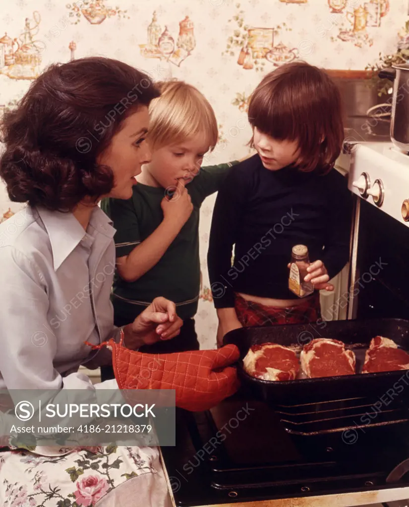 1970s WOMAN MOTHER 2 CHILDREN BOY GIRL KITCHEN OVEN DOOR OPEN CHECKING STEAKS MEAT ADDING SPICE COOKING