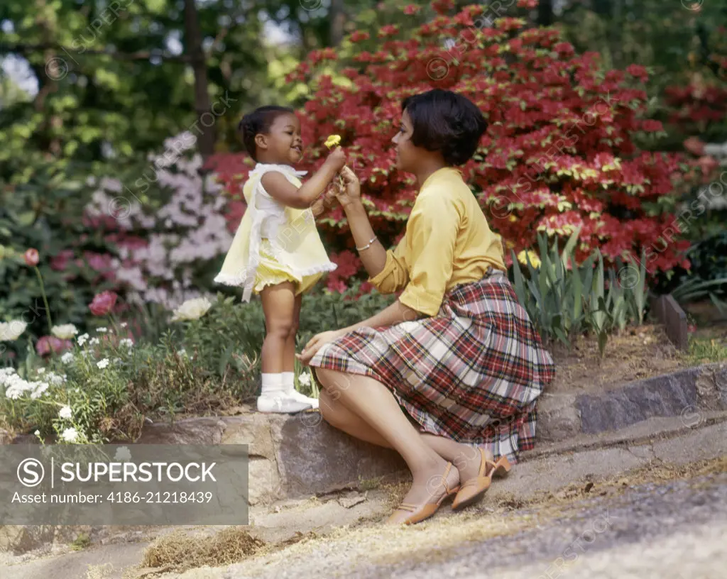 1970s SMILING AFRICAN AMERICAN WOMAN AND LITTLE GIRL OUTDOORS NEAR SPRING FLOWERS DAUGHTER GIVING YELLOW FLOWER TO MOTHER