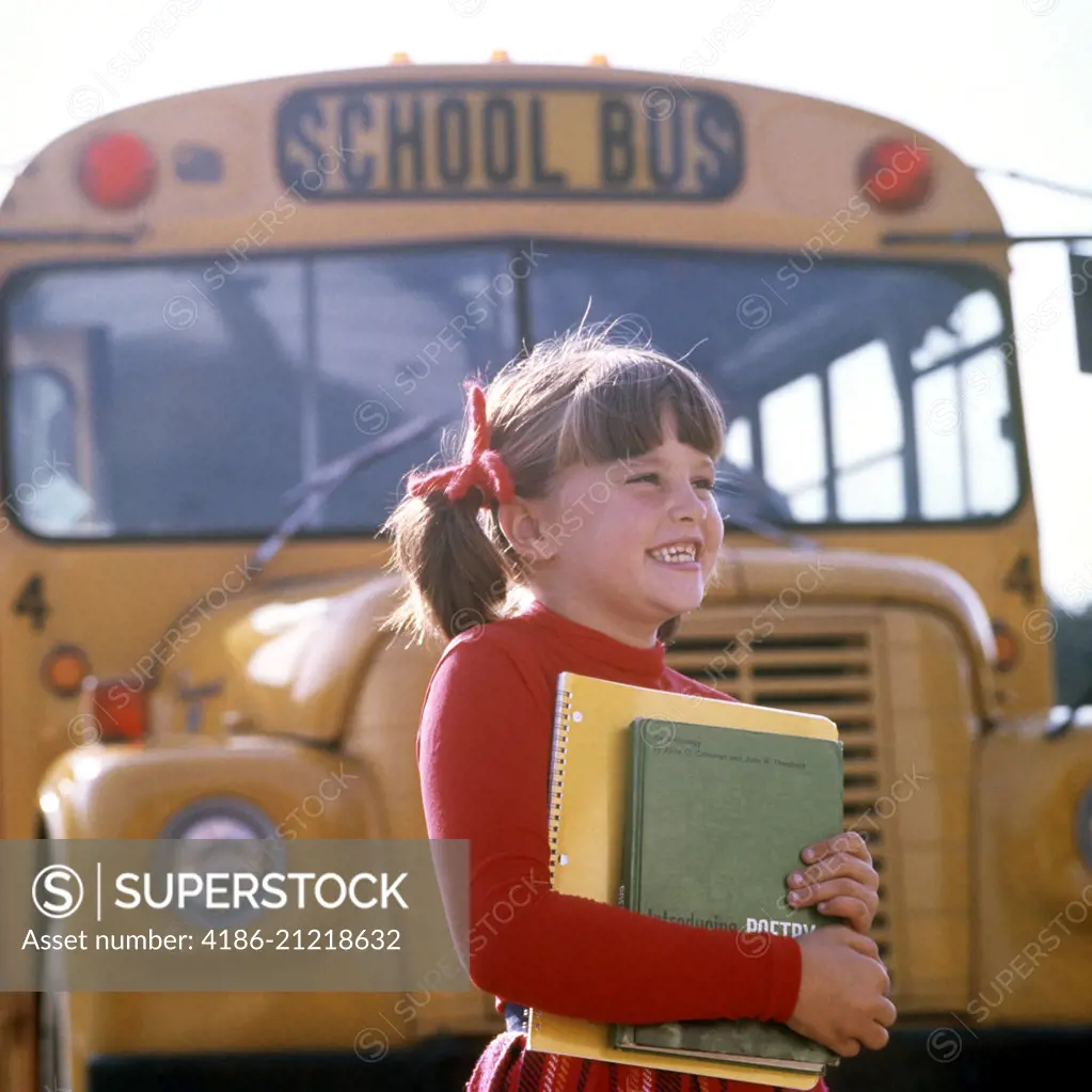 1970s SMILING ELEMENTARY SCHOOL GIRL STANDING FRONT OF BUS CARRYING BOOKS SMILING 
