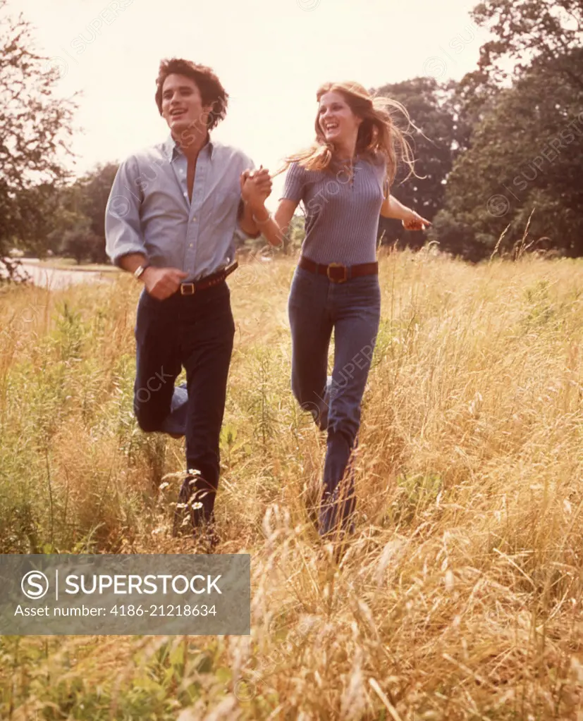 1970s SMILING HAPPY COUPLE RUNNING THROUGH MEADOW GRASS WEARING BLUE SHIRTS DENIM JEANS