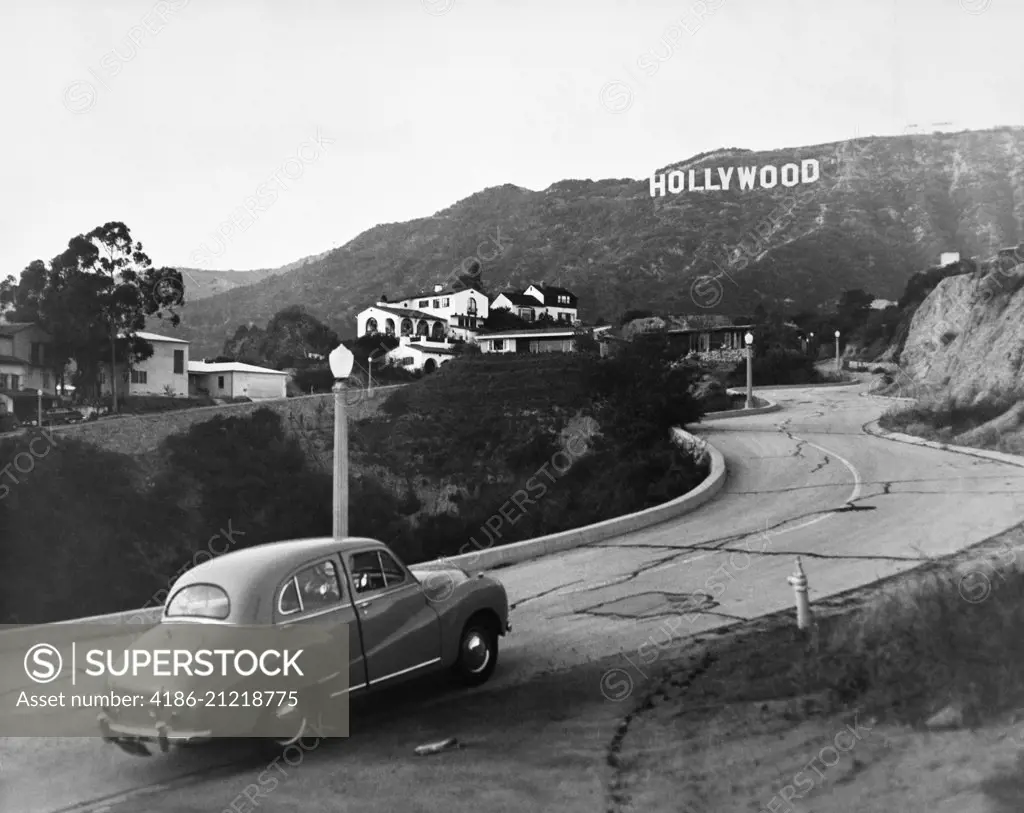 1950s AUSTIN CAR DRIVING UP THE HOLLYWOOD HILLS WITH HOLLYWOOD SIGN IN DISTANCE LOS ANGELES CA USA 
