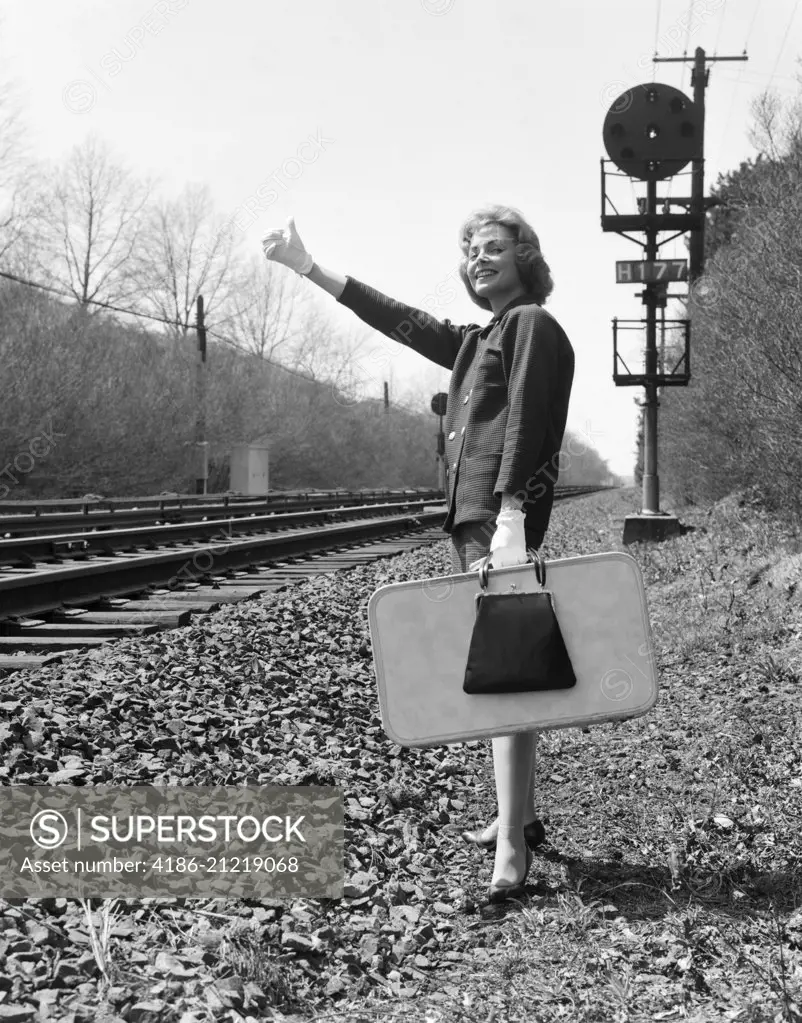 1950s 1960s WOMAN SMILING HOLDING SUITCASE STANDING BESIDE RAILROAD TRACKS GLOVED HAND THUMB IN THE AIR TO HITCH A RIDE