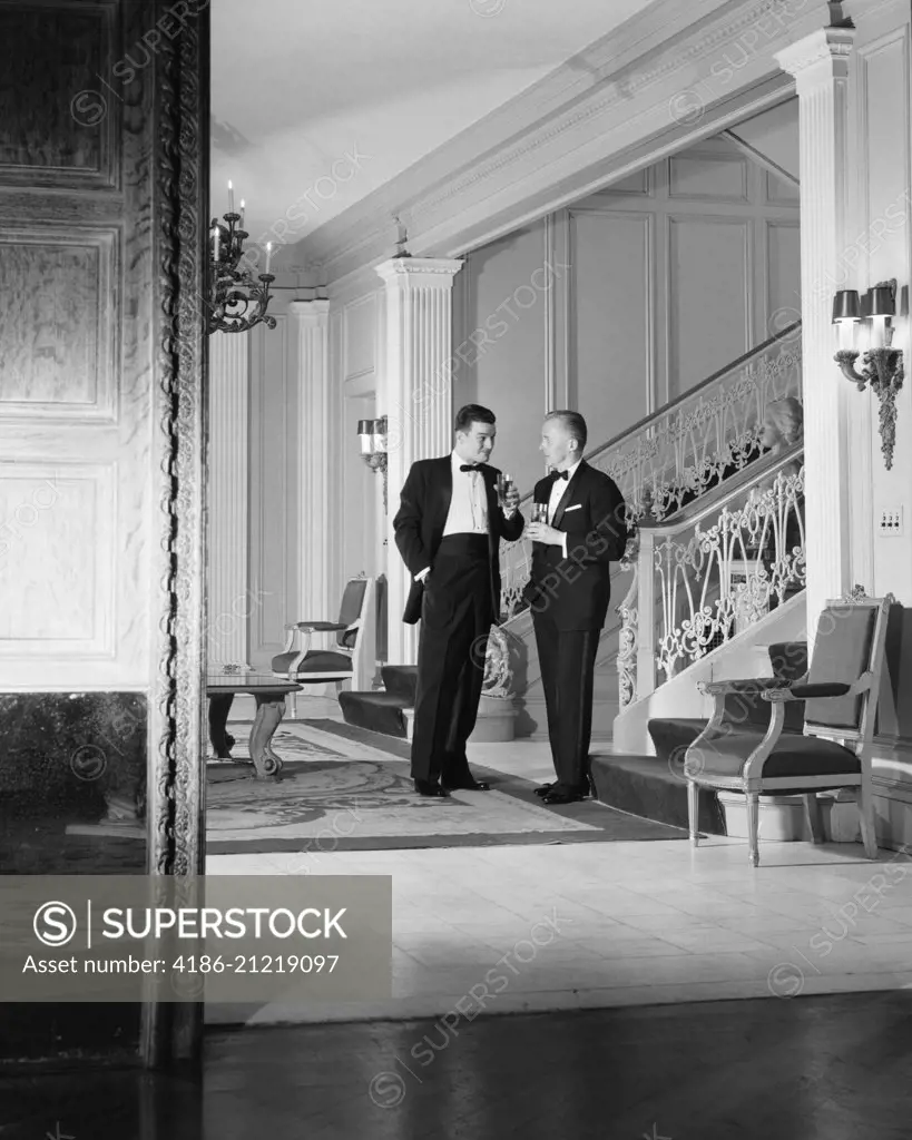 1950s PAIR OF MEN IN TUXEDOS HOLDING DRINKS AND TALKING SHOWN THROUGH DOORWAY OF LARGE ELEGANT HOME OR CLUB