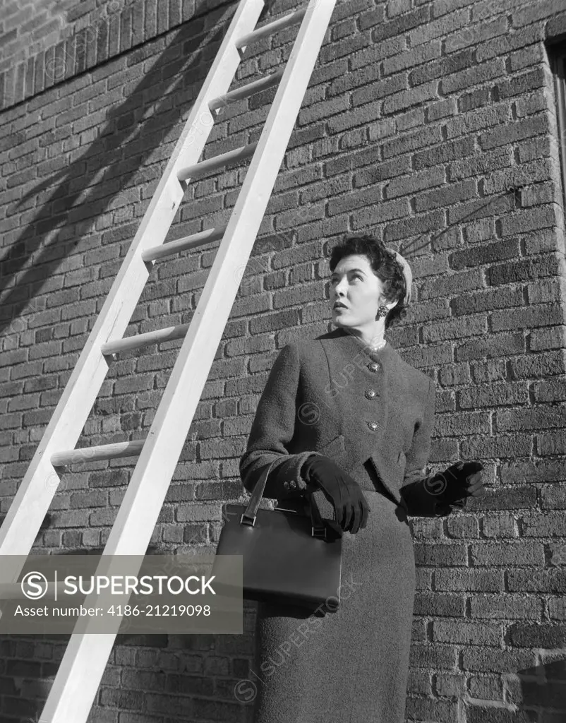 1950s PROVOKING BAD LUCK WOMAN WEARING SUIT AND HAT CARRYING HANDBAG WALKING UNDER LADDER