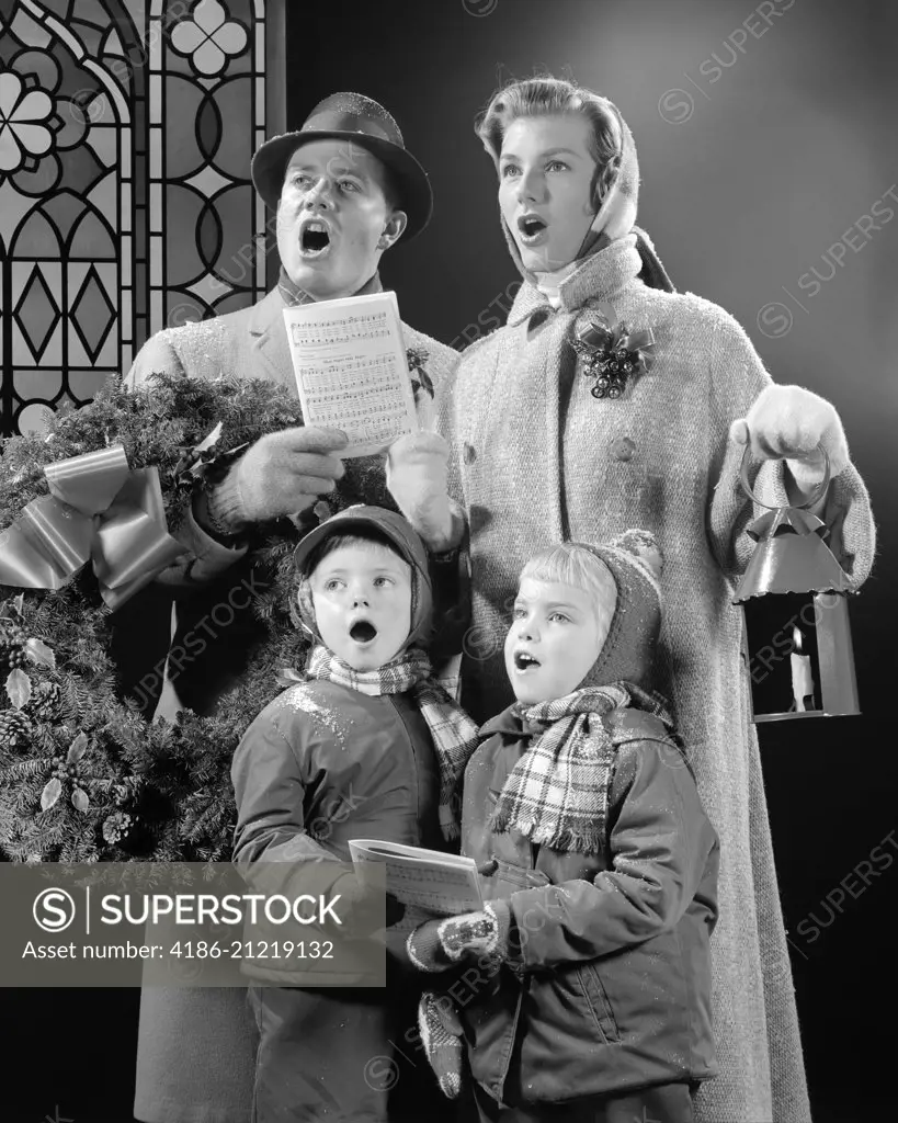 1950s FAMILY OF FOUR SON DAUGHTER SINGING CHRISTMAS CAROLS DAD HOLDING WREATH MOM HOLDING CANDLE LANTERN