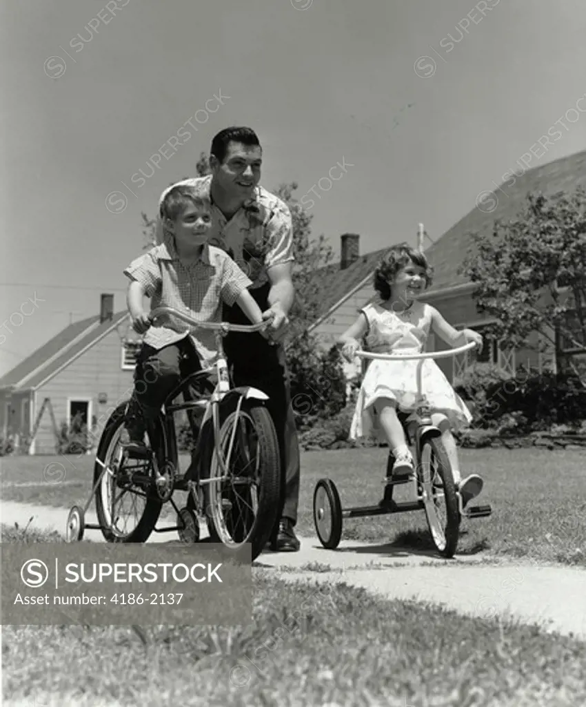 1950S Father Pushing Boy On Bike With Training Wheels And Girl On Tricycle Neighborhood Sidewalk