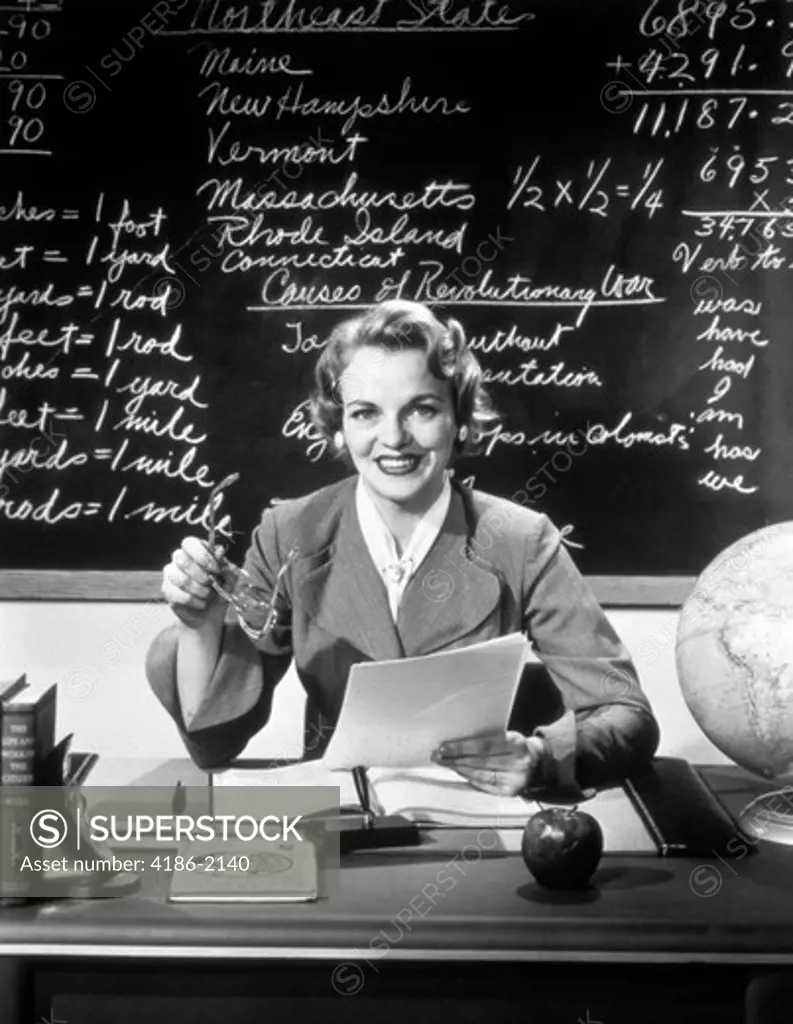 1950S Smiling Woman Elementary School Teacher Sitting Behind Desk In Front Of Blackboard Looking At Camera