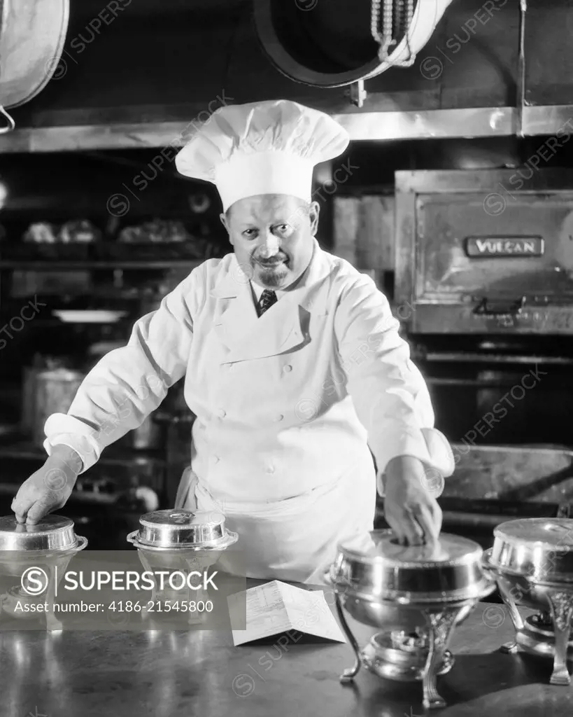 1920s 1930s SERIOUS FRENCH CHEF IN KITCHEN WITH SMALL SILVER CHAFING DISHES LOOKING AT CAMERA