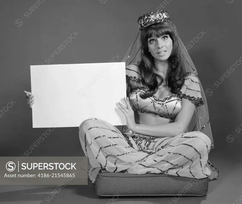 1960s WOMAN IN BELLY DANCE COSTUME SITTING ON CUSHION LOOKING AT CAMERA HOLDING BLANK SIGN
