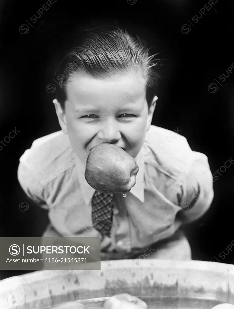 1930s BOY BOBBING FOR APPLES WITH AN APPLE IN HIS MOUTH LOOKING AT CAMERA