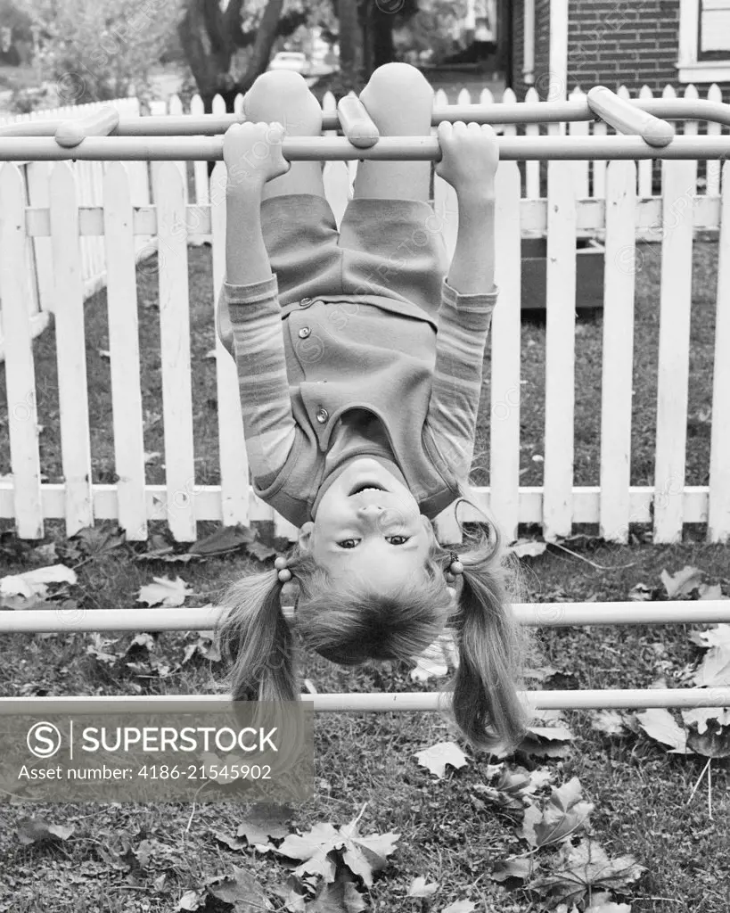 1970s SMILING YOUNG GIRL PIG TAILS HANGING UPSIDE DOWN ON BACKYARD JUNGLE GYM