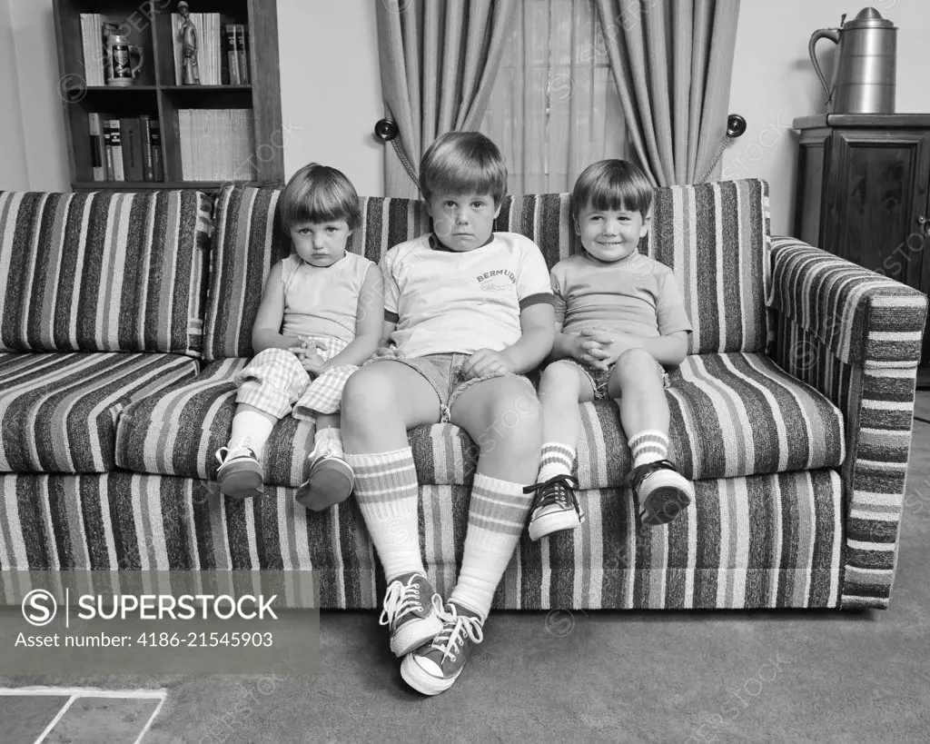 1970s THREE SIBLINGS SITTING ON COUCH LOOKING AT CAMERA