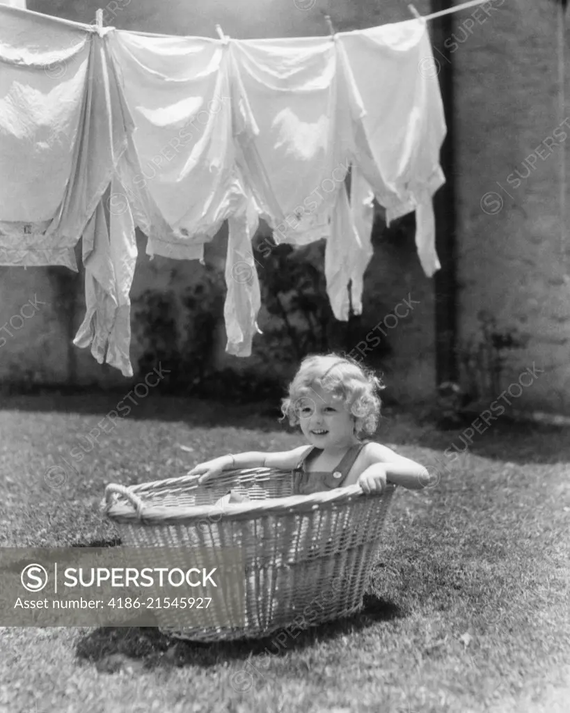 1930s 1940s GIRL OUTDOORS SITTING IN LAUNDRY BASKET UNDER CLOTHESLINE FULL OF SHIRTS