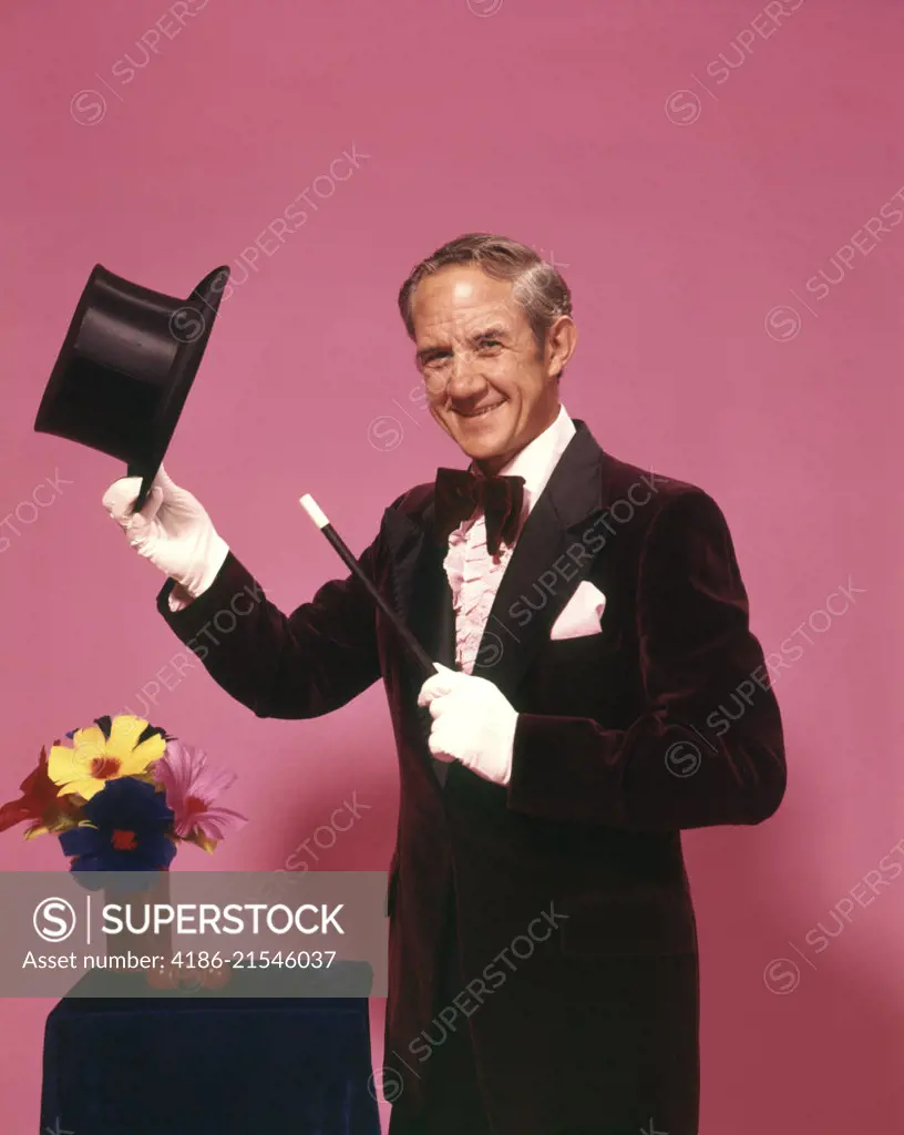 1970s MAN MAGICIAN LOOKING AT CAMERA TIPPING TOP HAT WITH WHITE GLOVES BOW TIE MAGIC WAND FLOWERS 