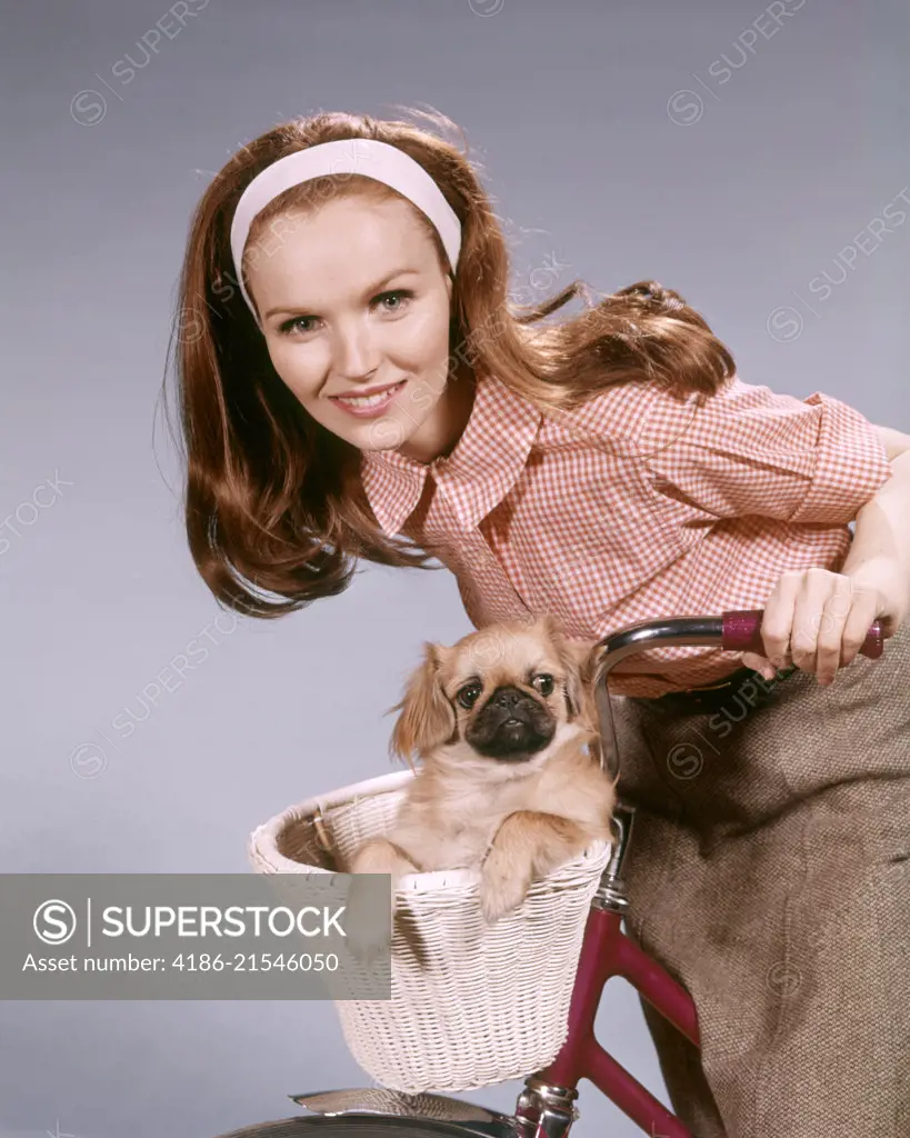 1960s SMILING WOMAN LOOKING AT CAMERA LEANING ON HANDLEBARS OF BICYCLE PET PUG DOG IN BASKET
