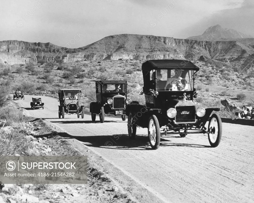 1960s LINE OF 1920s MODEL T AND 1930s MODEL A FORD AUTOMOBILES ON GRAVEL ROAD IN SOUTHWEST USA
