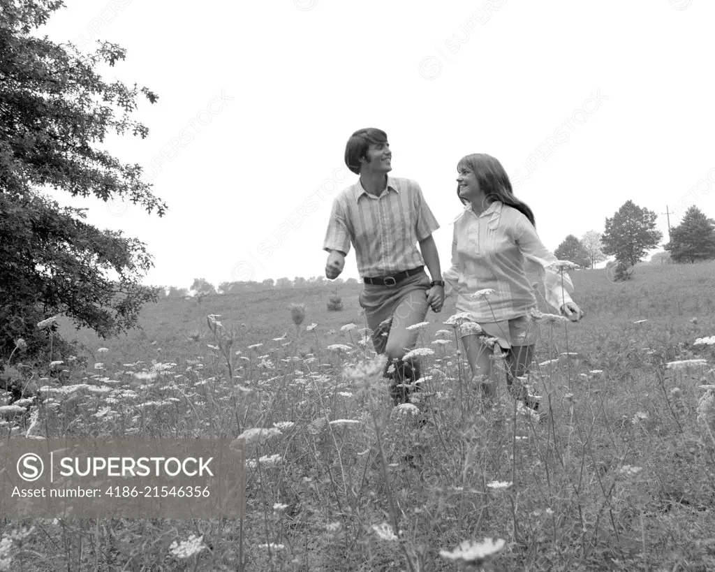 1970s COUPLE HOLDING HANDS WALKING IN HIGH FIELD WITH QUEEN ANNE'S LACE