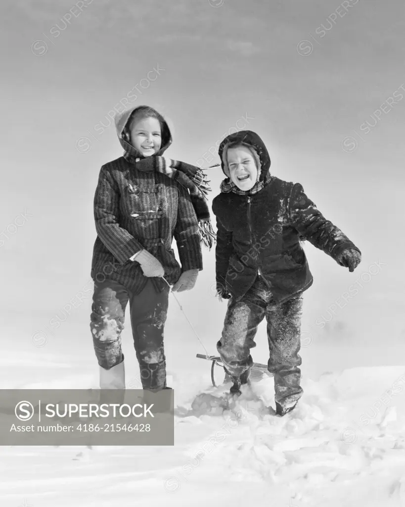 1950s 1960s SMILING BOY GIRL PULLING SLED IN WINTER SNOW LOOKING AT CAMERA