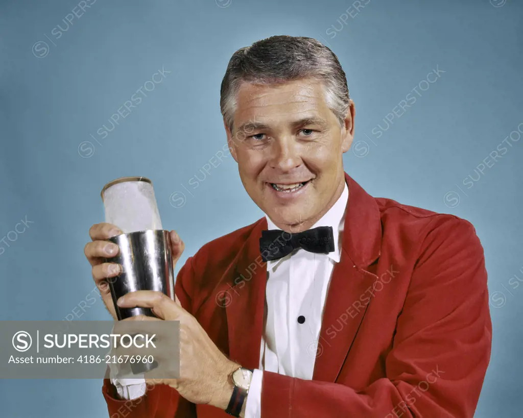 1960s 1970s SMILING MAN BARTENDER IN RED JACKET LOOKING AT CAMERA HOLDING COCKTAIL SHAKER MAKING MIXED DRINK
