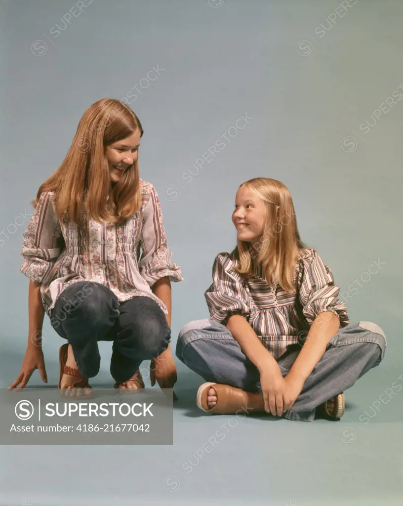 1970s TWO TEEN GIRLS SISTERS WEARING BLUE JEANS DENIM PEASANT SHIRTS SITTING AND KNEELING SMILING LOOKING AT EACH OTHER 