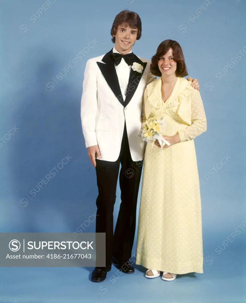 1970s TEEN COUPLE DRESSED FOR PROM GIRL YELLOW LONG DRESS GOWN BOY WHITE TUXEDO BLACK LAPELS STANDING TOGETHER LOOKING AT CAMERA