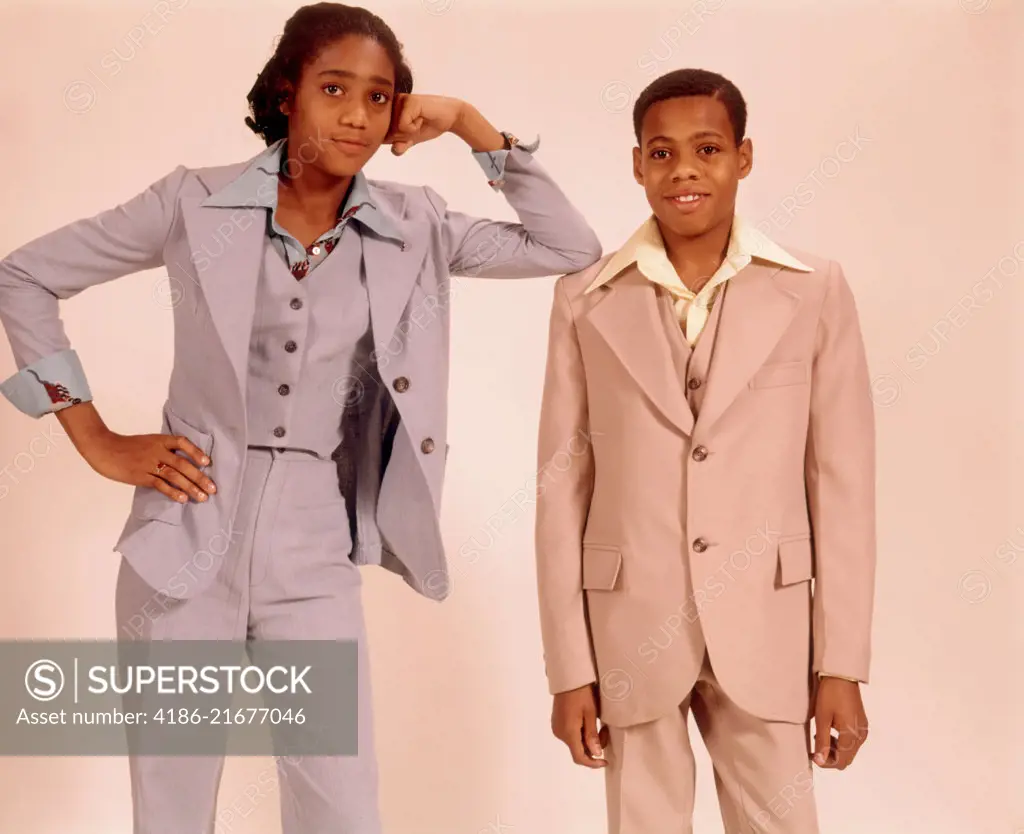 1970s TWO AFRICAN AMERICAN TEENAGERS A BOY IN A BEIGE SUIT AND GIRL IN A BLUE PANTS SUIT STANDING TOGETHER LOOKING AT CAMERA