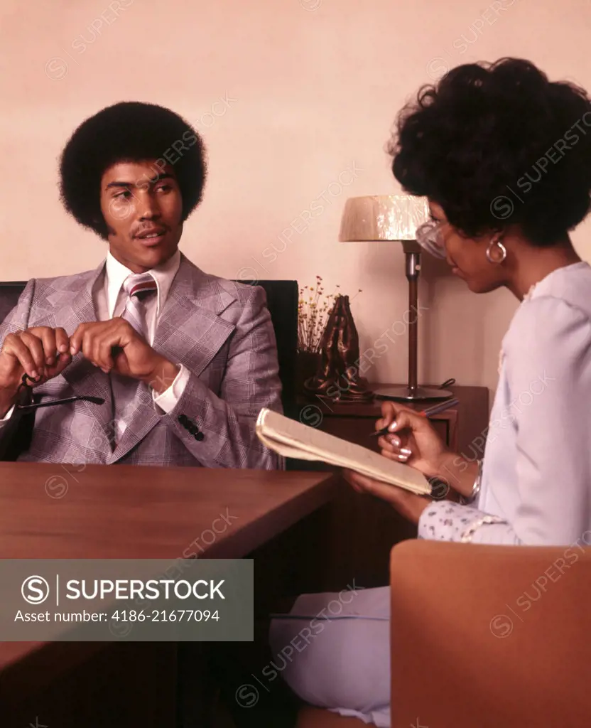 1970s AFRICAN AMERICAN STENOGRAPHER TAKING SHORTHAND DICTATION FROM AFRICAN AMERICAN BUSINESSMAN SEATED AT OFFICE DESK