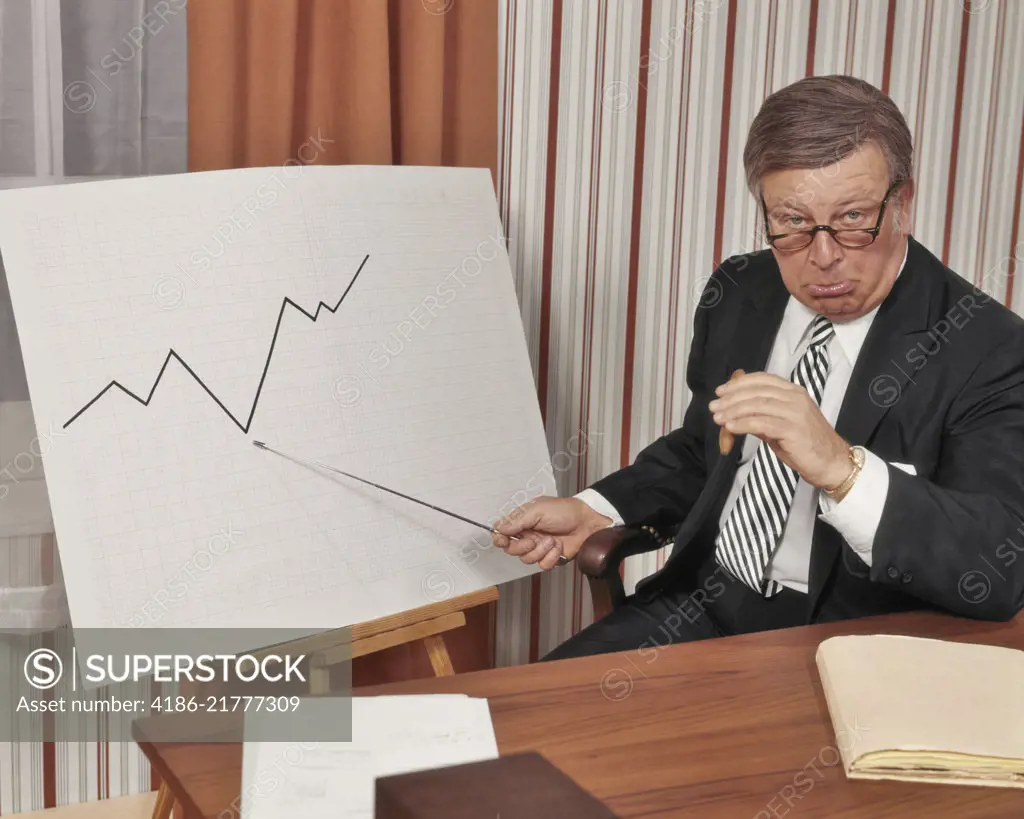 1970s UNHAPPY BUSINESS MAN LOOKING AT CAMERA POINTING TO GRAPH CHART INDICATING A DECLINE IN SALES PROFITS BUSINESS