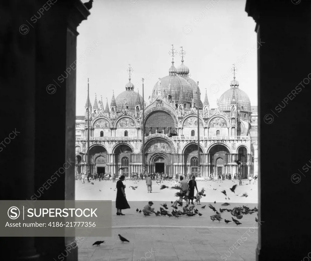 1920s 1930s VIEW THROUGH DOORWAY TO PEOPLE FEEDING PIGEONS IN FRONT OF ST. MARKS CATHEDRAL VENICE ITALY