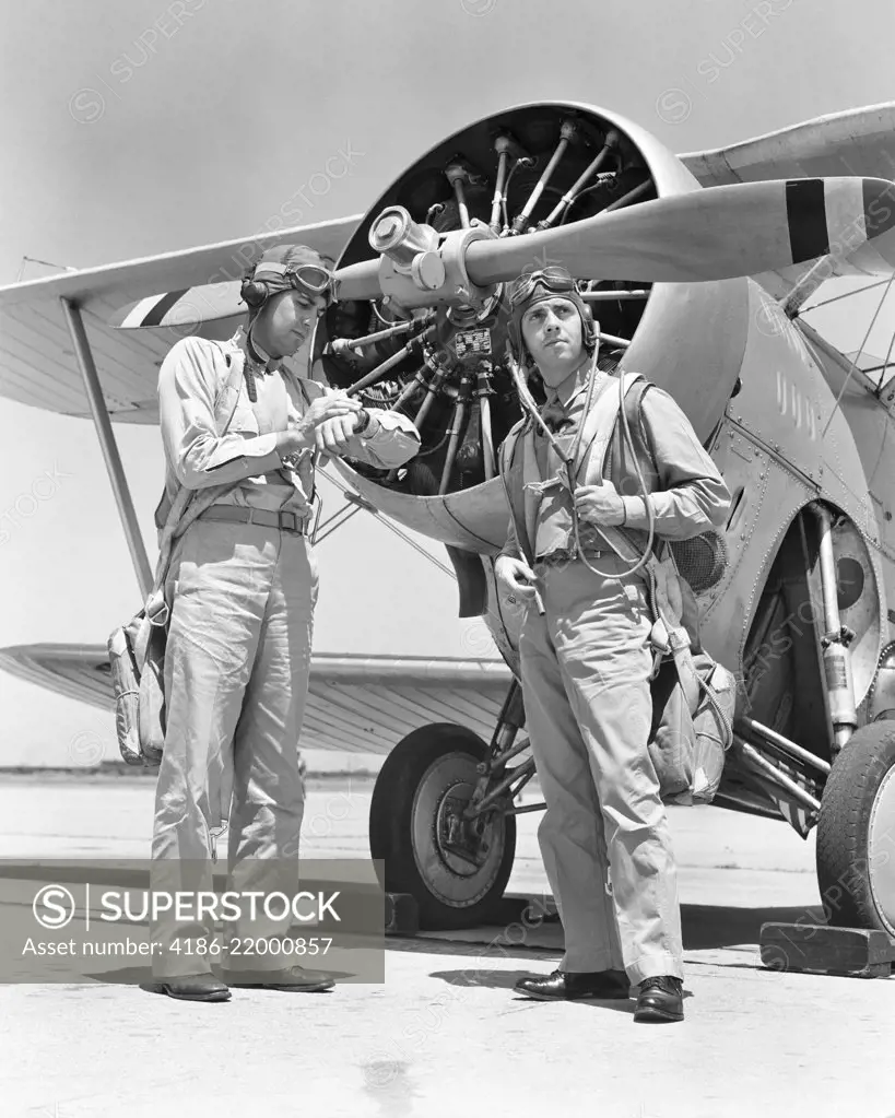 1940s TWO US NAVY PILOTS WEARING LEATHER FLYING HELMETS AND PARACHUTES STANDING READY BY PROPELLER BIPLANE