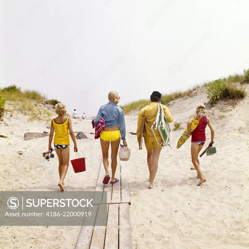 1970s FAMILY MOTHER FATHER TWO BOYS WALKING ON TO BEACH CARRYING CHAIRS UMBRELLA GEAR 
