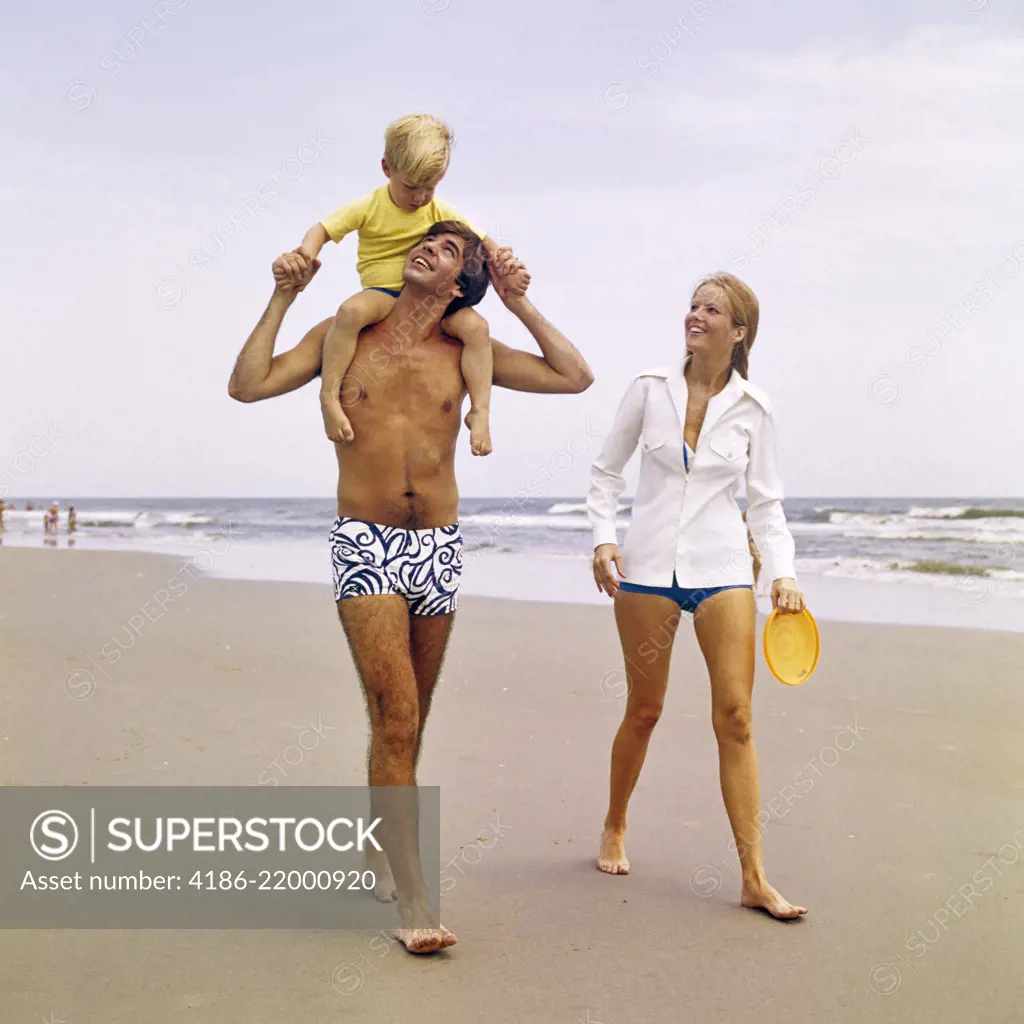 1970s YOUNG FAMILY WALKING DOWN BEACH TODDLER RIDING ON FATHERS SHOULDERS