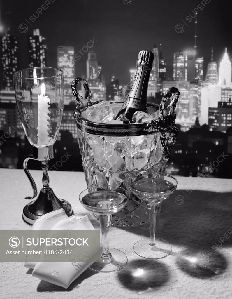 Champagne Bottle In Ice Bucket With Glasses Candle & Napkins On Table & New York City Night Skyline In Background
