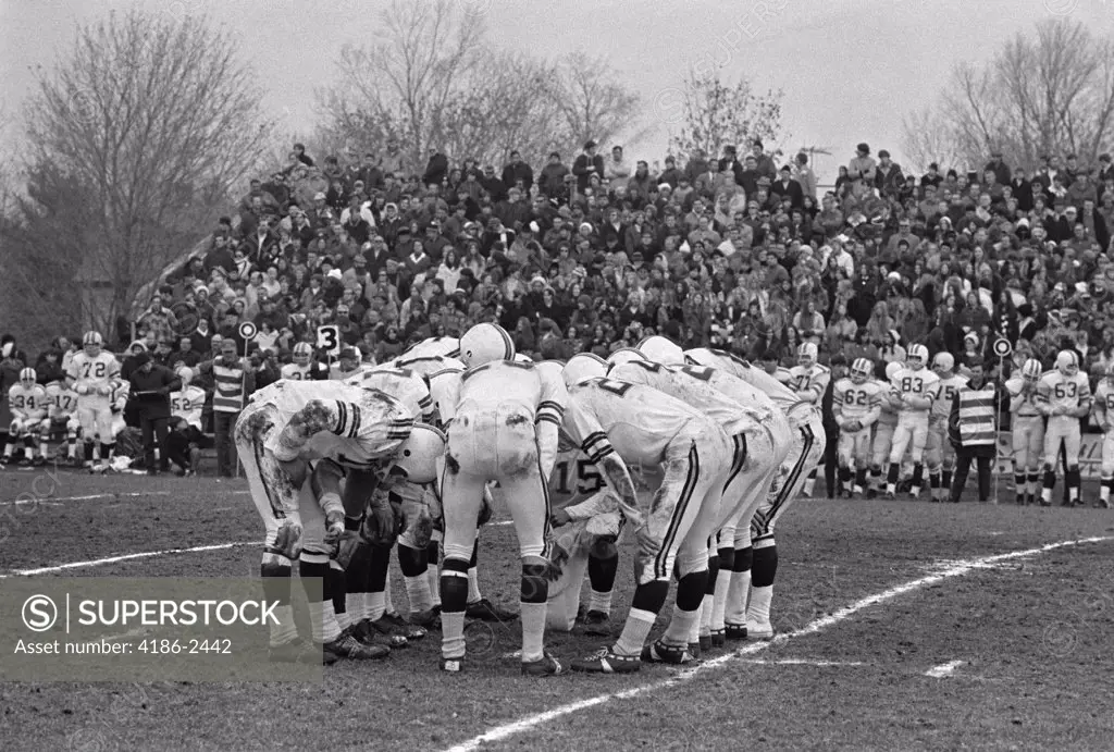 1980S Football Huddle On Playing Field With Spectators In Bleachers In The Background Outside