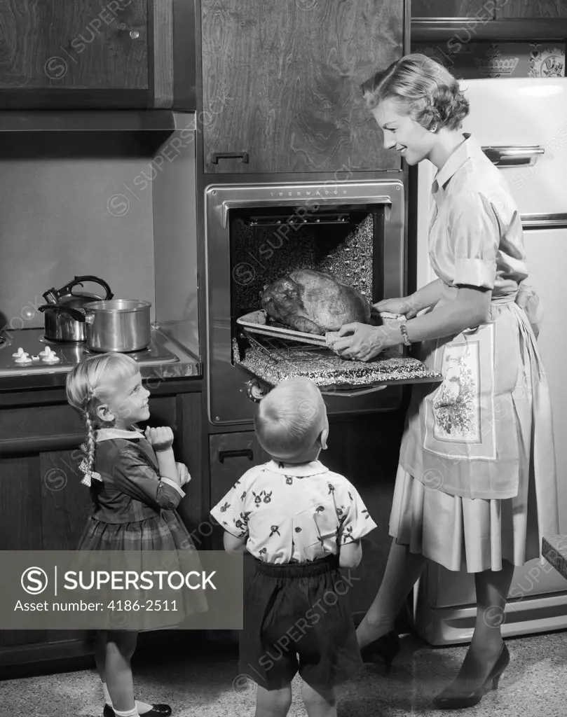1950S Daughter And Son Watching Mother Remove Roast Thanksgiving Turkey Dinner From Electric Kitchen Oven