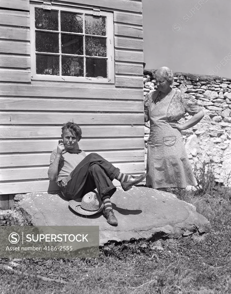 1940S Farm Boy Leaning Against House Smoking Pipe With Grandmother Looking Around Corner At Him With Hand On Hip