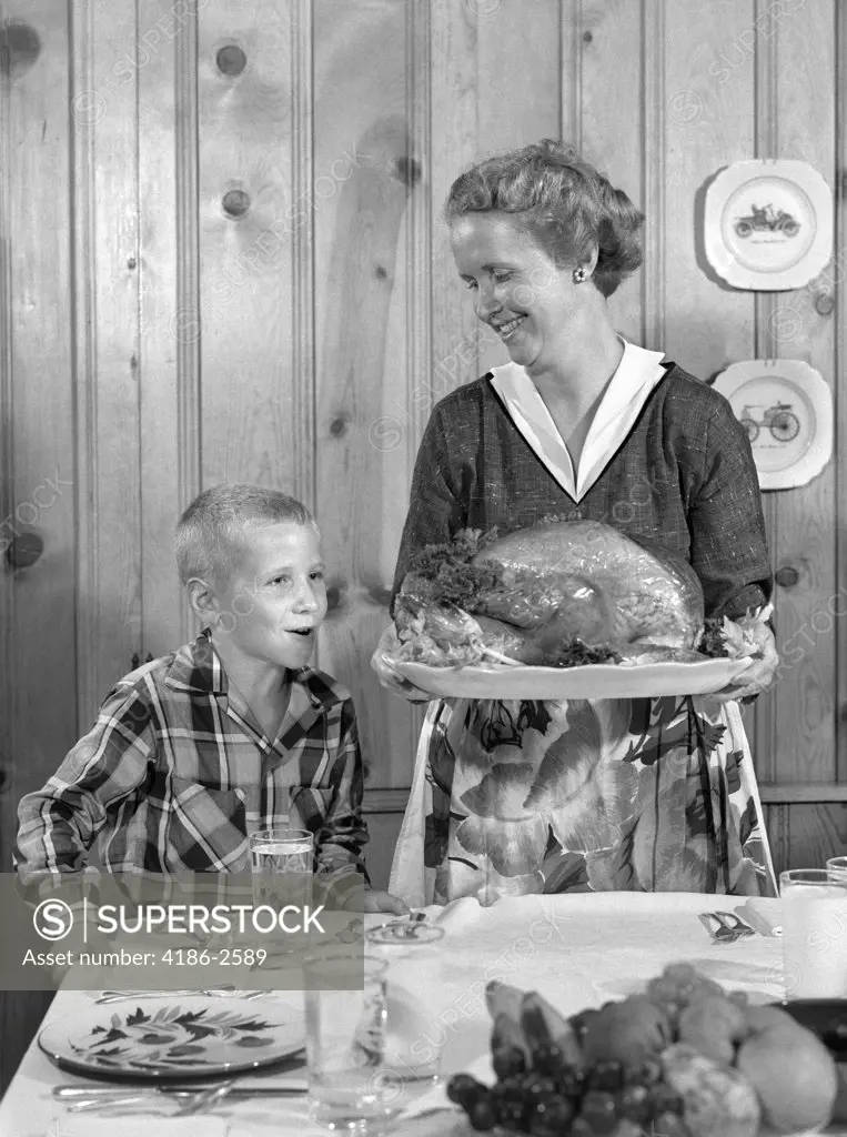 1950S Woman In Apron Putting Turkey On Table With Son Looking On Eagerly