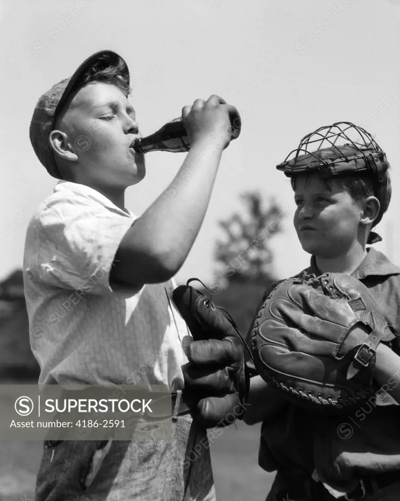 1930S Pair Of Boys Wearing Baseball Gloves One With Catcher'S Mask Watching Other Wearing Cap Drink Bottle Of Soda