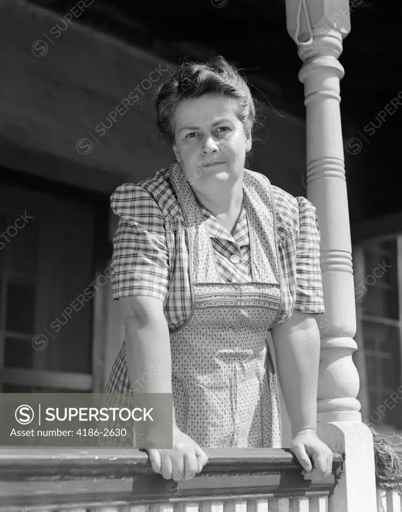 1940S Older Woman In Plaid Dress & Apron Leaning On Porch Railing Of Farmhouse