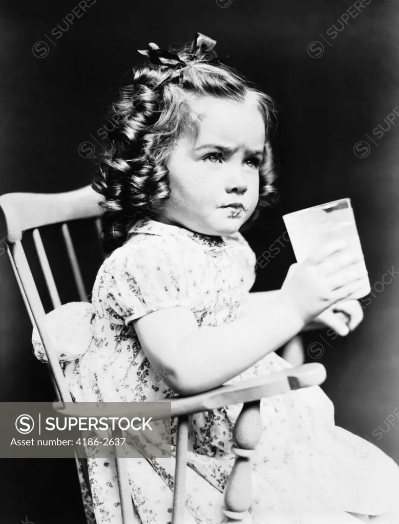 1930S Child Girl Sitting In High Chair Holding Glass Of Milk Serious Look Bow In Hair Baloney Curls Print Dress Toddler