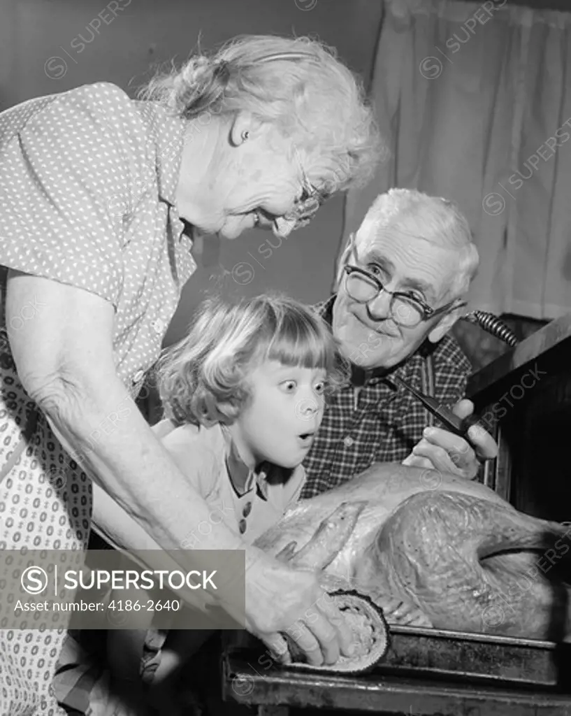 1950S 1960S Excited Little Girl Granddaughter With Grandparents Watching Cooked Turkey Coming Out Of Oven